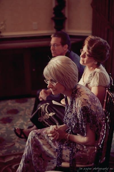 A group of three older adults sits closely together indoors. The two women wear floral dresses, and the man is in a suit. They appear attentive and engaged, with soft lighting highlighting their expressions.