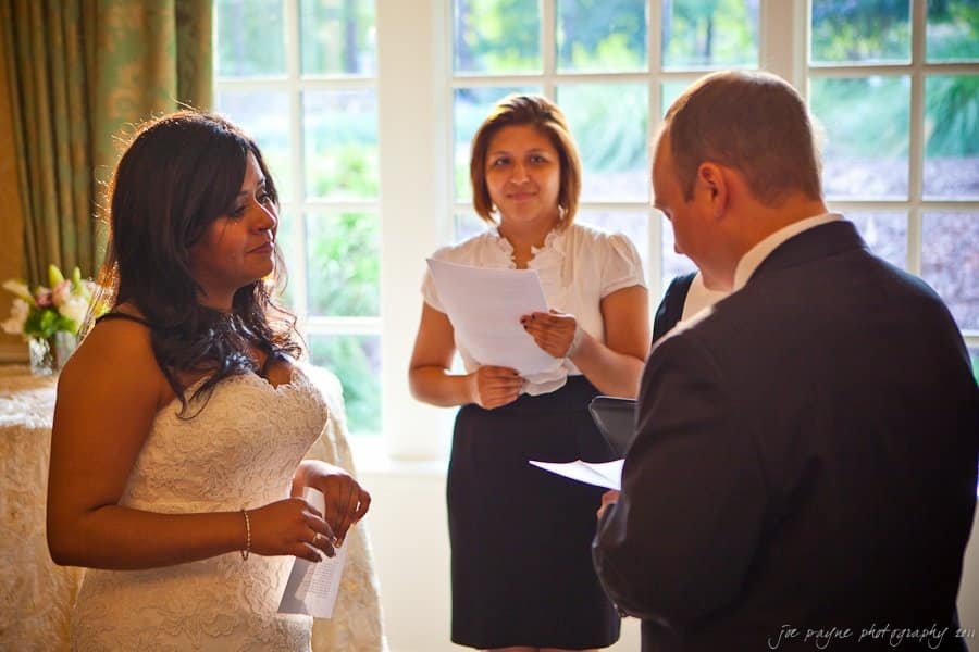 A bride and groom stand facing each other, exchanging vows in a sunlit room with large windows. A woman stands between them, holding papers and smiling. The bride wears a lace dress, and the groom is in a dark suit.
