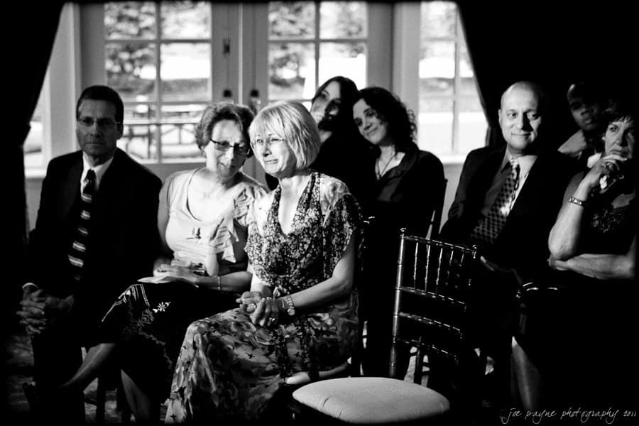 A black and white photo of a group of people seated indoors, attentively watching something. The setting has large windows in the background, and the people are dressed in semi-formal attire.