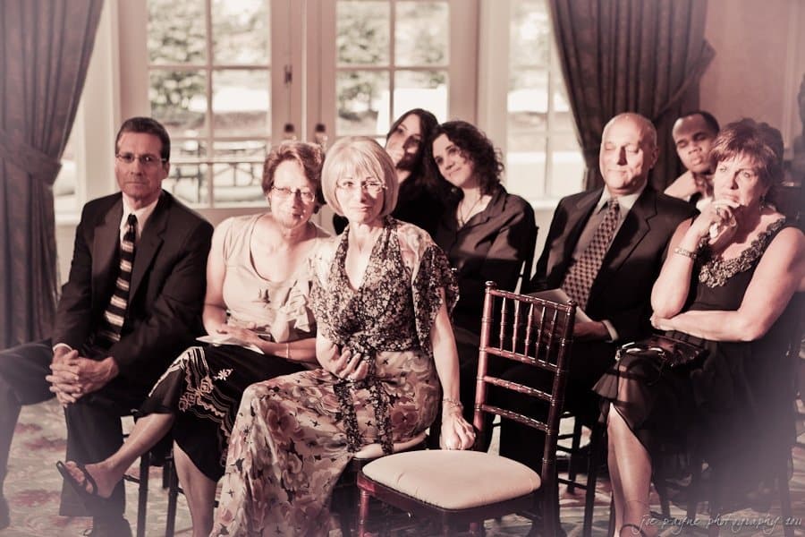 A group of people in formal attire are seated indoors, facing the camera. The setting appears to be a formal event with curtains and large windows in the background. The mood is casual, with some individuals smiling.