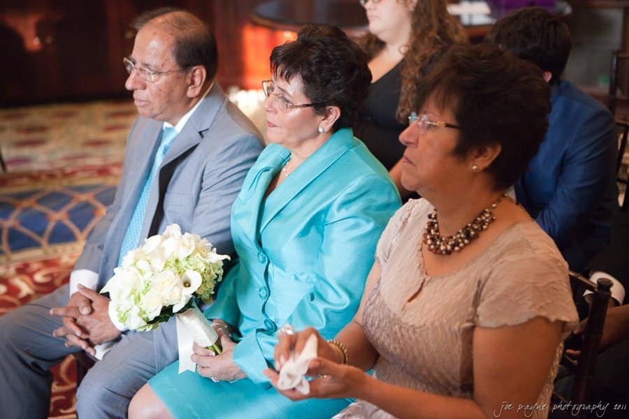 Three seated older adults at an indoor event. A man in a light gray suit and blue tie sits beside two women; one in a bright turquoise suit holding a bouquet, and the other in a light beige dress. They appear engaged in the proceedings.