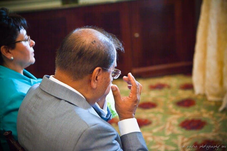 A man in a suit wipes his glasses while sitting next to a woman in turquoise clothing. They are seated indoors on patterned carpet with wood-paneled walls in the background.