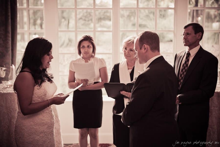 A bride and groom stand facing each other, exchanging vows in a brightly lit room with a large window behind them. Three people, two women and one man, stand nearby, witnessing the ceremony.
