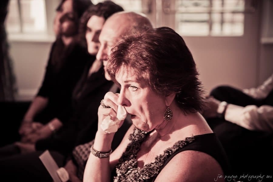 A woman in a patterned dress sits indoors, wiping tears with a tissue. Other people in formal attire are seated beside her, blurred in the background. The setting appears emotional and reflective.