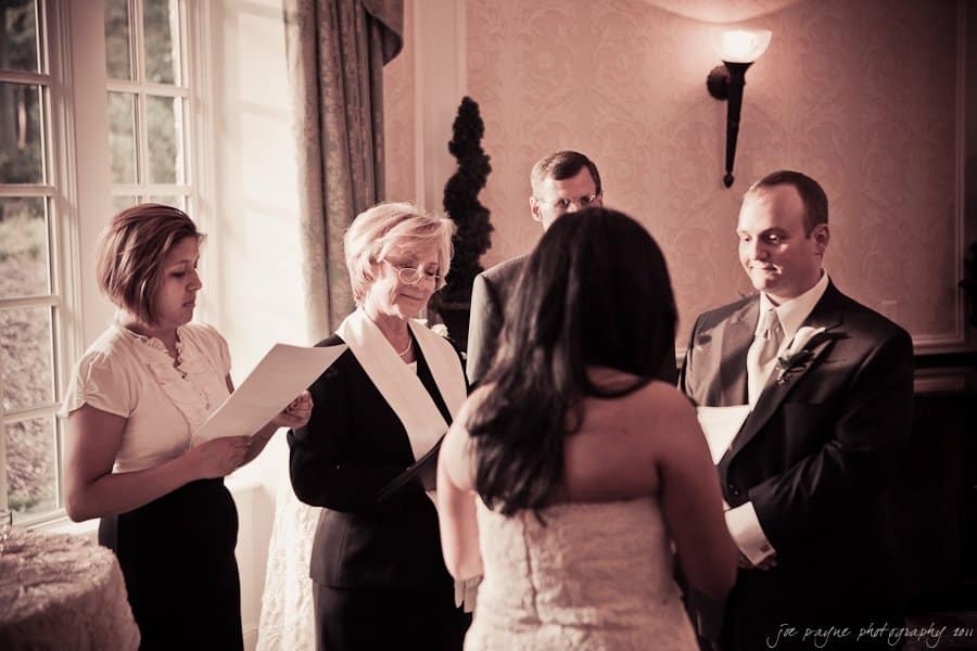 A sepia-toned image of a small wedding ceremony. A couple stands facing an officiant holding a paper, with another person to the officiants side. Two men are in suits. The setting is indoors with ornate walls and a large window.