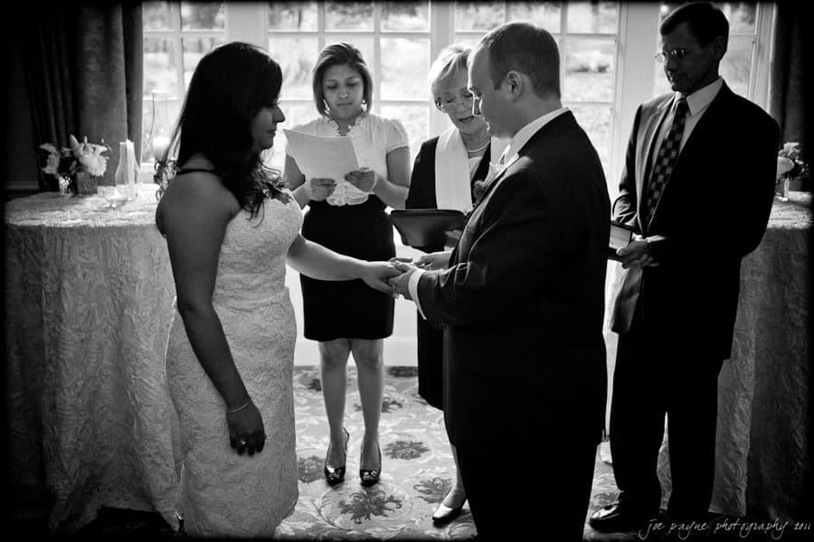 A bride and groom stand during a wedding ceremony, exchanging rings. Two women officiate, holding papers, while another man looks on. The scene is set in a room with large windows and flowers on a table.