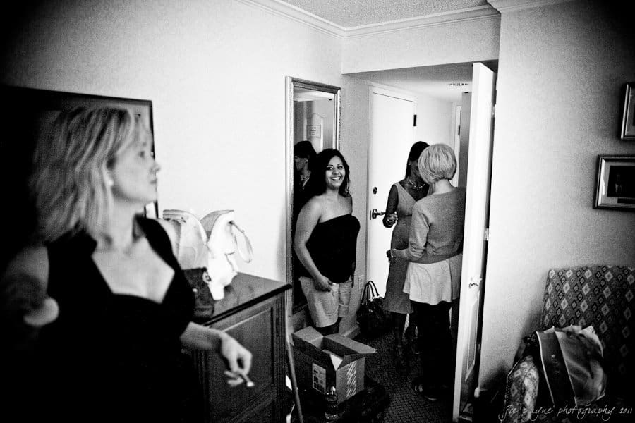 A group of women in a room preparing for an event. One woman in focus is smiling, while another stands in the foreground. The room is cluttered with bags and boxes. Its a black-and-white photo with a casual, candid atmosphere.