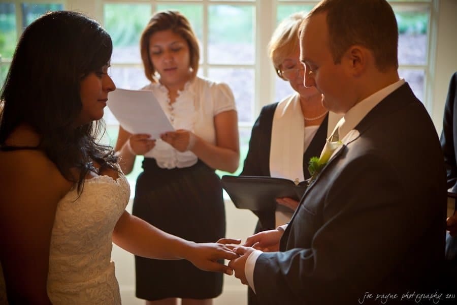 A bride and groom exchange rings during a wedding ceremony. The groom places a ring on the brides finger while two women stand in the background, one reading from a script. A large window lets in natural light behind them.