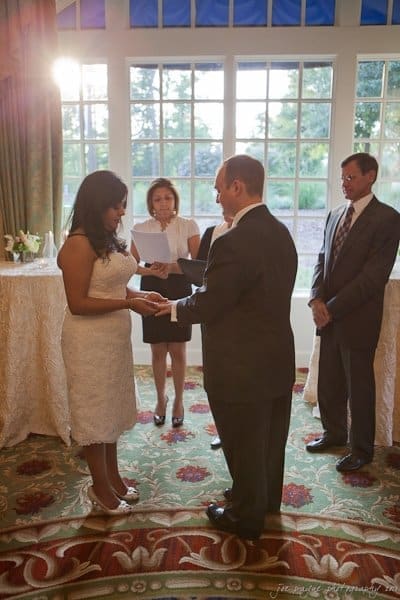 A couple is exchanging vows indoors with a windowed backdrop. The bride is in a white dress, and the groom wears a dark suit. Two guests stand nearby, one holding paper, seemingly officiating the ceremony, while sunlight streams in.