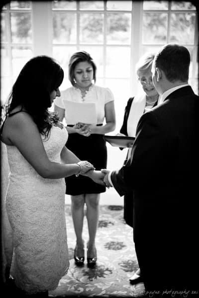 A bride and groom stand facing each other, holding hands, during a wedding ceremony. Two officiants stand nearby; one is reading from a paper. The scene is indoors with a large window in the background. The photo is in black and white.