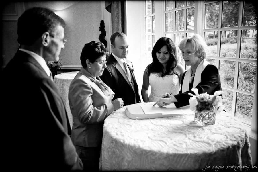 A wedding ceremony scene with five people gathered around a table by a window. A woman in a light suit gestures toward a document, while a bride and groom look on, alongside two other individuals. The room is elegantly decorated.