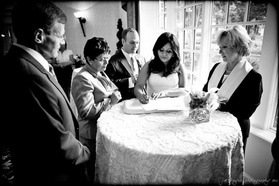 A black and white photo of a wedding ceremony. A bride is signing a document on a table surrounded by family members and an officiant. They are in a lit room with a large window in the background.