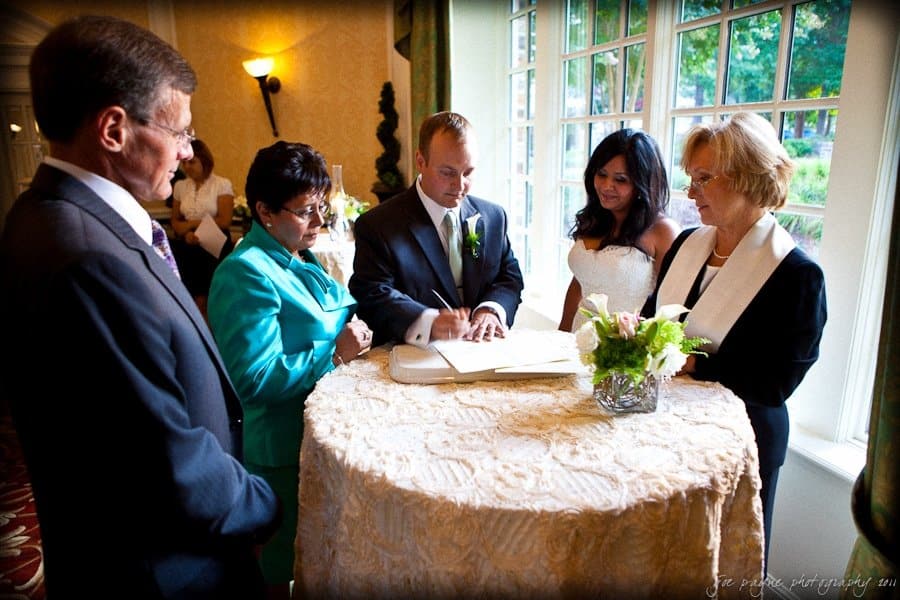 A couple signs their marriage certificate on a round table covered with a lace cloth. Two officials stand nearby while another woman looks on. The room is warmly lit, with a vase of flowers and large windows in the background.