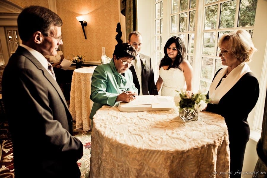 A wedding ceremony inside a decorated room. Five people surround a table with a guest book. A woman in green signs the book, while others watch. One person is in a wedding dress. Flowers adorn the table, and sunlight streams through the windows.