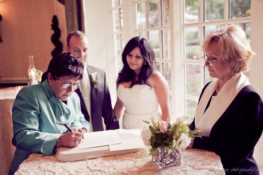 A wedding ceremony scene with a couple next to an officiant and a woman signing a book. The couple stands near a table with floral decorations. The room is softly lit, with large windows in the background showing greenery.