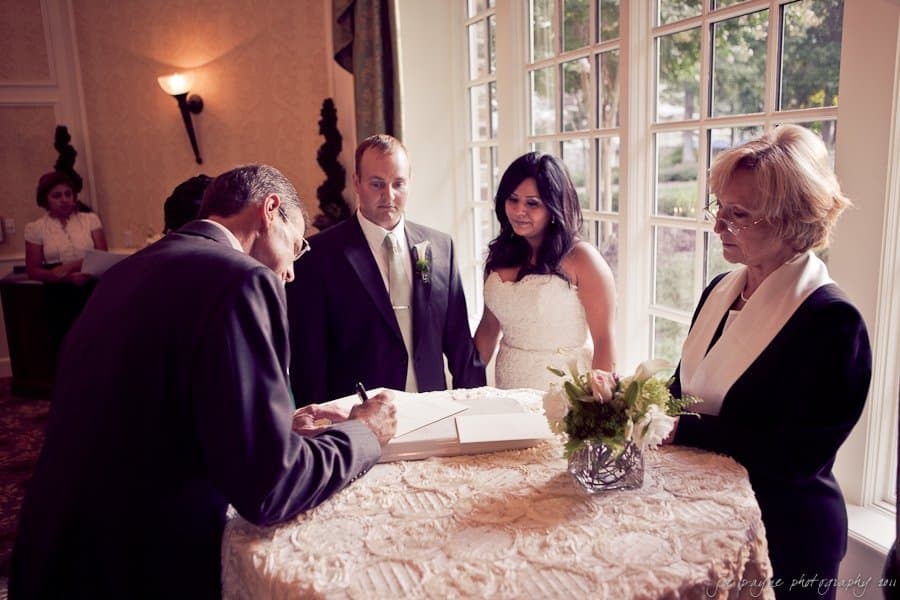 A wedding ceremony scene where a man signs a document at a round table. The bride, groom, and two other individuals stand nearby. The room is softly lit, and a window offers a view of greenery outside.