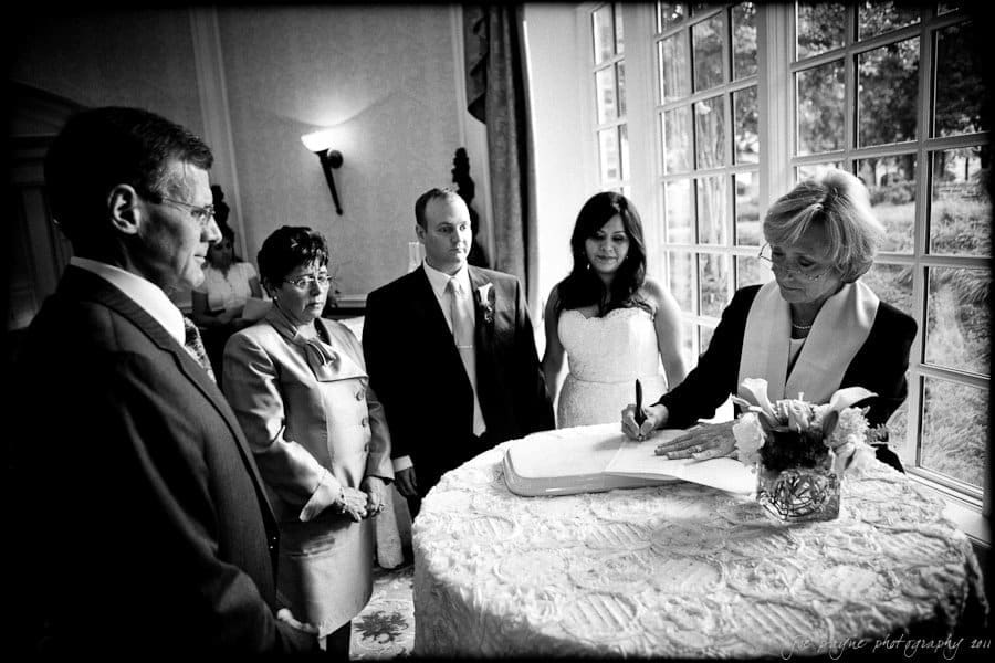 A black-and-white photo of a wedding ceremony. A man and woman stand across a table from another couple, while a woman writes in a book. The table is covered with a lace cloth and a flower arrangement. Large windows reveal a garden view outside.