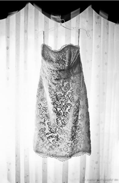 A strapless, lace-embroidered dress hangs on a hanger in front of sheer curtains. The dress features intricate floral designs and delicate scalloped edges, with light filtering softly through the fabric.
