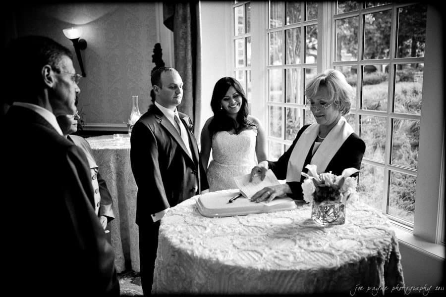A black-and-white image of a wedding ceremony. A bride and groom stand beside an officiant signing documents on a round, lace-covered table. Two other people look on. Large windows reveal a lush garden outside.