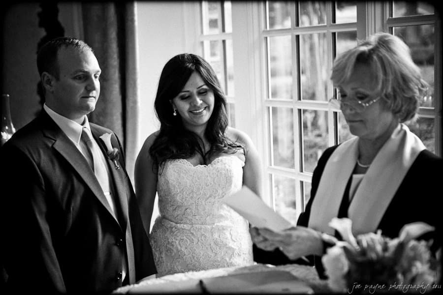 A couple stands before an officiant during a wedding ceremony. The bride, in a lace gown, smiles while the groom gazes forward. The officiant, holding a document, stands beside a window with natural light filtering through. Black and white image.