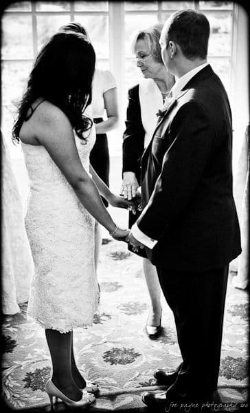 A bride and groom stand holding hands during a wedding ceremony. They are facing each other, with the officiant standing between them. The scene is indoors with light streaming through a large window behind them.