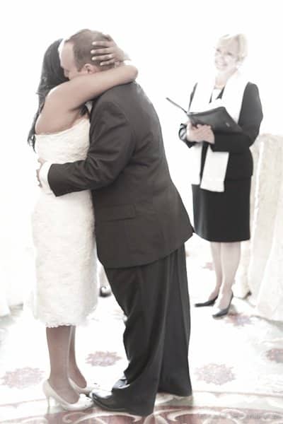 A couple in formal attire embraces during a wedding ceremony. The bride wears a white dress and the groom is in a dark suit. An officiant with a book and scarf stands nearby, smiling. The room is bright and softly lit.