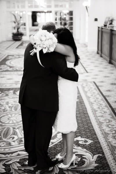 A couple embraces in a warmly lit hallway with patterned carpet. The woman holds a bouquet of white flowers as they share a close moment. The scene exudes love and intimacy.