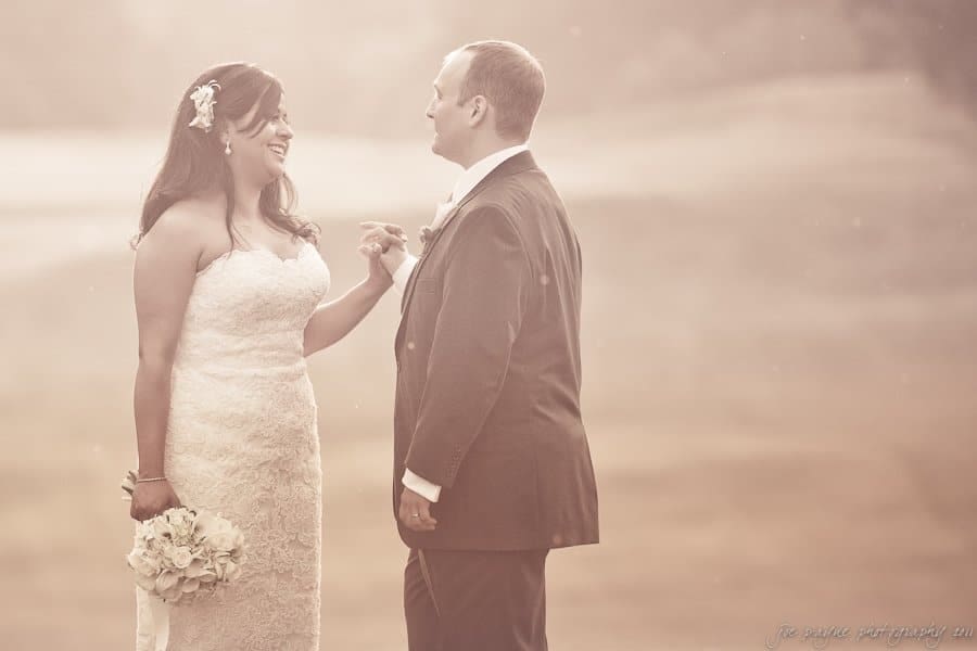 A bride and groom stand facing each other outdoors, holding hands. The bride wears a lace gown and the groom is in a suit. The background is softly blurred, creating a dreamy, romantic atmosphere.