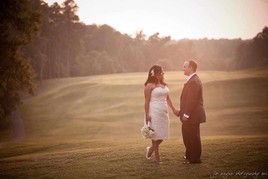 A bride and groom stand hand in hand on a grassy field during sunset. The bride wears a white dress and holds a bouquet, while the groom is in a dark suit. Trees line the horizon in the background.