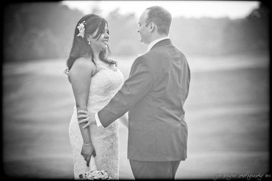 A bride and groom stand smiling at each other. The bride holds a bouquet and wears a lace dress with a flower in her hair. The groom is in a suit. They are outdoors on a misty day, with soft lighting creating a romantic atmosphere.