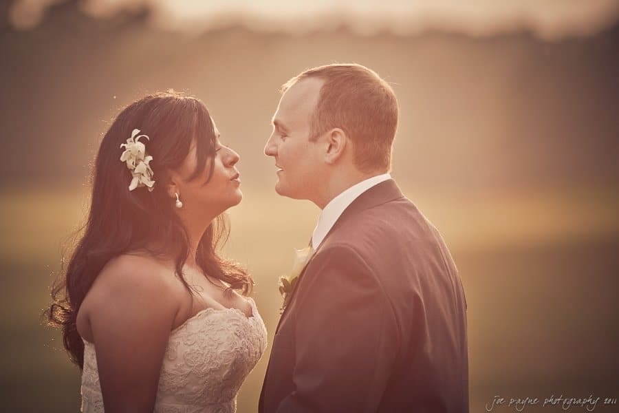 A bride and groom stand close, gazing into each others eyes during sunset. The bride wears a white dress with a flower in her hair, and the groom is in a suit with a boutonniere. The background is softly lit with a warm glow.
