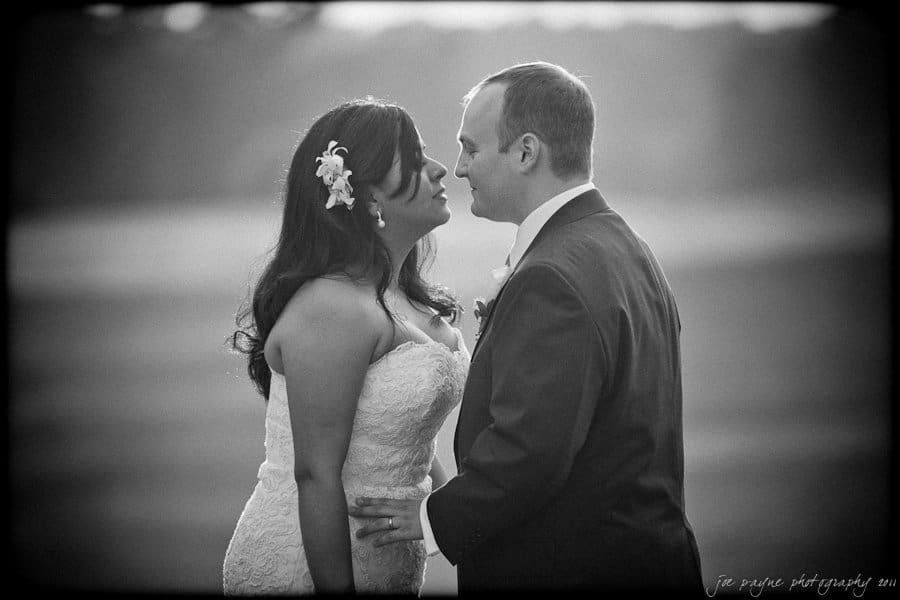 A couple dressed in formal wedding attire stands closely facing each other. The bride wears a strapless gown with flowers in her hair, while the groom wears a suit. The background is softly blurred, creating a romantic atmosphere.