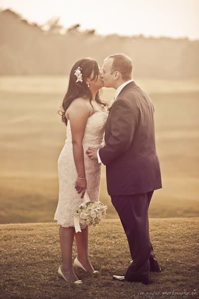 A bride and groom share a kiss outdoors on a grassy field. The bride wears a white lace dress and holds a bouquet. The groom is dressed in a suit. The background is softly lit with a warm, golden hue.