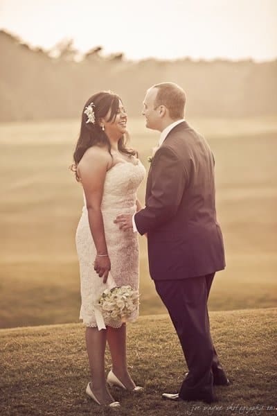 A bride and groom stand together on a grassy field, with the sun setting behind them. The bride holds a bouquet of flowers and wears a lace dress, while the groom is in a suit. They are smiling at each other, creating a romantic atmosphere.