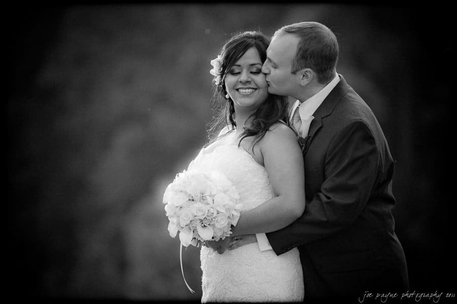 A groom lovingly kisses the cheek of a smiling bride holding a bouquet of white flowers. The couple is dressed in wedding attire against a blurred outdoor background.