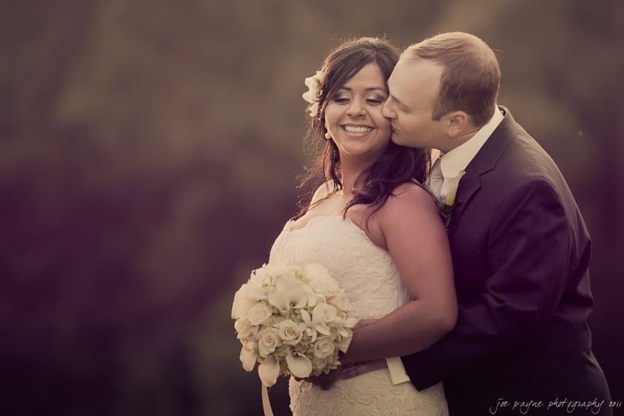 A couple in formal wedding attire is embracing outdoors. The groom is kissing the brides cheek while she holds a bouquet of white flowers and smiles. The background is softly blurred, giving a romantic vibe.
