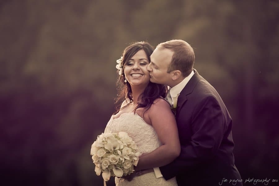 A groom kisses the smiling bride on the cheek as they pose together outdoors. She holds a bouquet of white roses. The background is softly blurred.