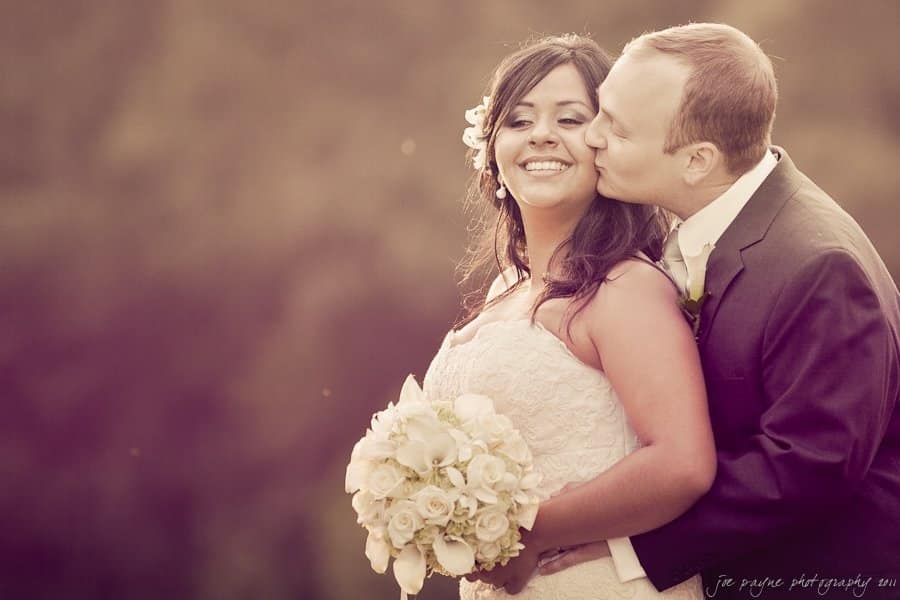A bride holding a bouquet of white flowers smiles while a groom kisses her cheek. They are outdoors, with a blurred background and warm lighting creating a romantic atmosphere.