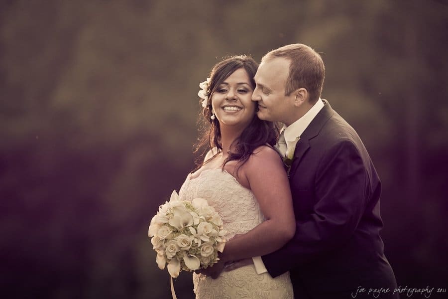 A bride and groom stand closely together outdoors. The bride holds a bouquet of white flowers and smiles, while the groom leans in affectionately. The background is a blurred, natural setting.