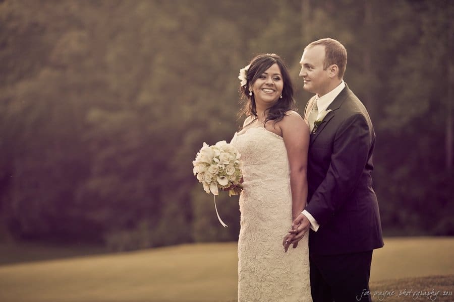 A bride and groom stand together in an outdoor setting. The bride is wearing a white dress and holding a bouquet, while the groom wears a dark suit. They are smiling and looking off-camera, with a blurred natural background behind them.