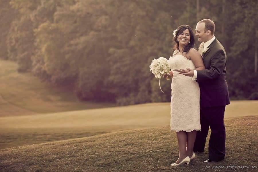 A smiling couple stands on a grassy hill during a wedding. The woman, in a white lace dress, holds a bouquet of white flowers. The man, in a dark suit, stands behind her. Trees and a soft, warm light fill the background.