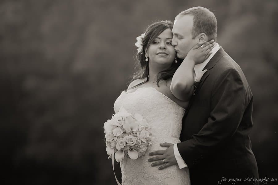 A bride and groom embrace in an outdoor setting. The bride holds a bouquet of white flowers, while the groom kisses her cheek. Both wear formal wedding attire. The background is blurred, emphasizing the couples intimate moment.