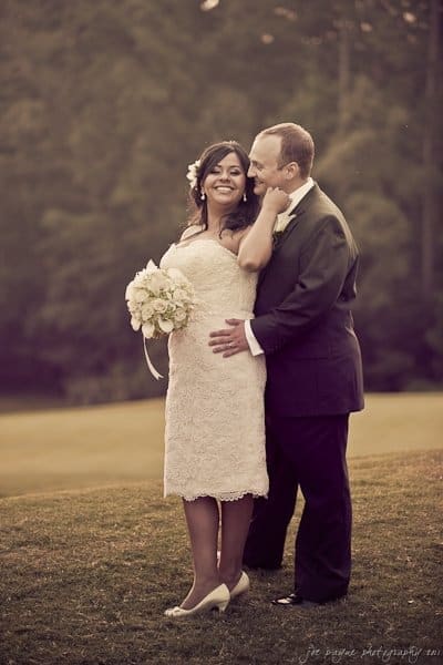 A bride and groom stand together outdoors. The bride, wearing a white dress and holding a bouquet, smiles broadly. The groom, in a dark suit, gently kisses her forehead. They are surrounded by greenery in a serene setting.
