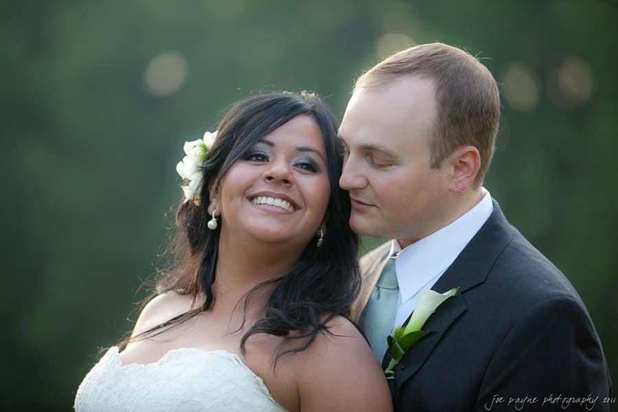 A bride and groom embrace outdoors, smiling at each other. The bride wears a white dress with a floral hairpiece, while the groom is in a dark suit with a white shirt and tie. The background is blurred green foliage.