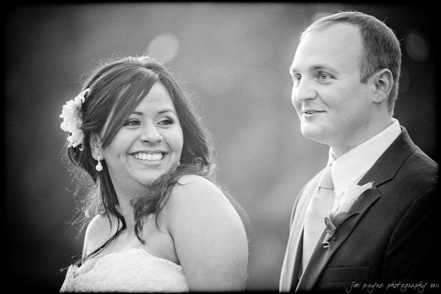 A black and white photo of a smiling couple. The woman, wearing a strapless dress and floral hair accessory, looks happy. The man, in a suit, gazes to the side. The background is softly blurred.
