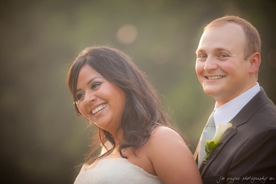 A bride and groom stand together outdoors, smiling warmly. The bride is in a white wedding dress, and the groom is in a suit with a white boutonniere. The background is softly blurred, giving a romantic feel.