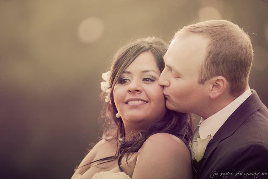 A couple embraces in soft, warm light. The man gently kisses the woman on the cheek as she smiles softly. The background is blurred, creating an intimate and tender atmosphere.