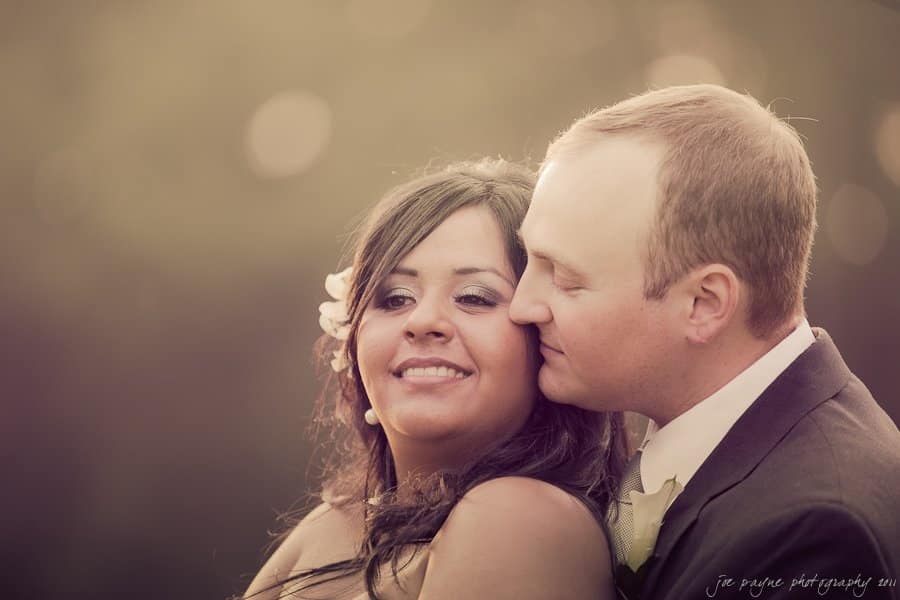 A couple shares an intimate moment outdoors. The woman smiles gently while leaning slightly back, and the man, wearing a suit, rests his head close to hers. The background is softly blurred with warm, golden lighting.