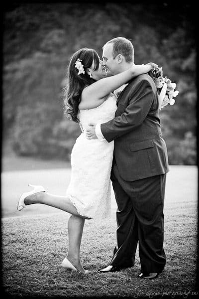 A bride and groom share a joyful embrace outdoors. The bride, in a knee-length dress and heels, lifts one foot off the ground, holding a bouquet. The groom, in a suit, holds her close. The background is a blurred natural setting. Black and white photo.
