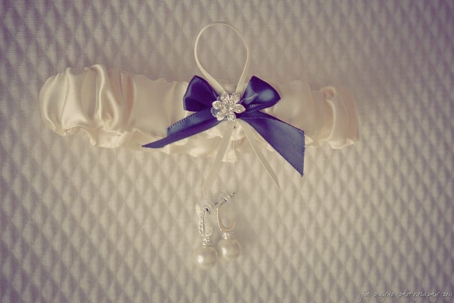 A white satin garter with a navy bow and pearl accents, adorned with a small flower brooch, is displayed on a textured white background. A pair of pearl earrings is attached to the garter.